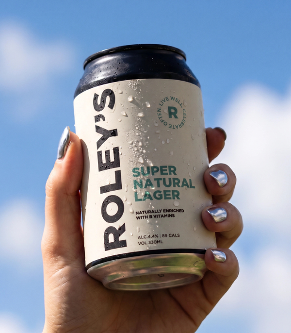 A hand with silver nail polish holds a can of Roley's Super Natural Lager against a bright blue sky with clouds.