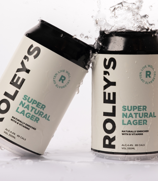 Two cans of Roley's Super Natural Lager beer, one lying on its side and one standing upright, both with water droplets splashing off them against a white background.